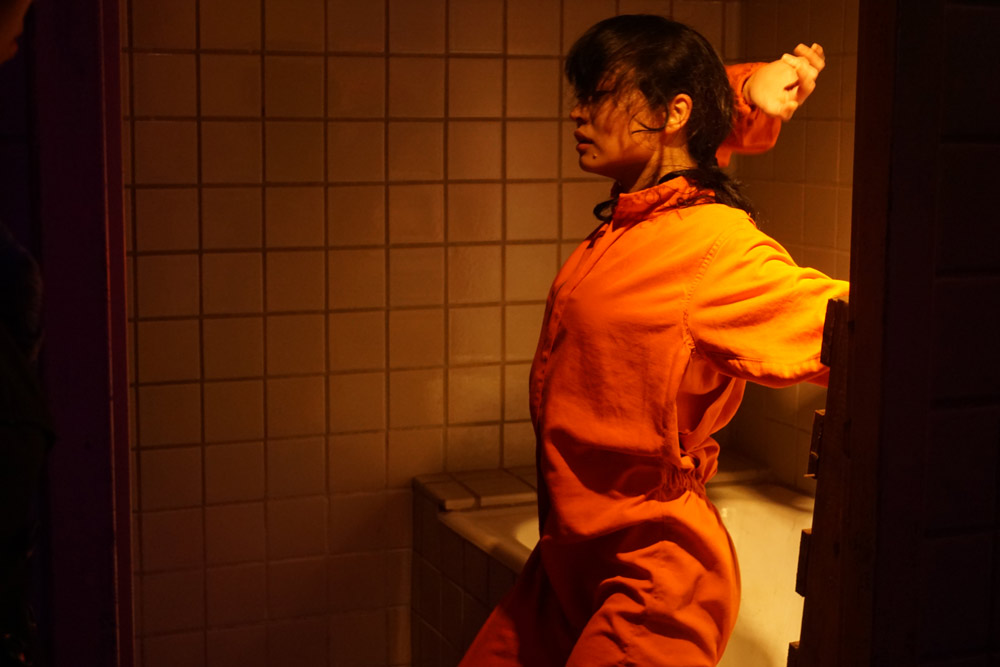 Japanese woman sitting at the edge of a bathtub, profile with her facing to left of screen. She is wearing an orange jumper and her left arm extends to her side and off the frame and her right hand is above her head.