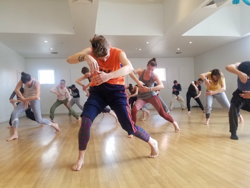 Woman standing in front of large group behind her leading a movement threading her right hand through a space that her left hand has created. The left hand is moving down and this picture captures the moment it is about to meet the right hand, which is extended in front of her chest. The students behind her are mimicking the movement. The teachers is dressed in a bright orange shirt and navy blue exercsie pants with orange pants down the side. Her legs are spread apart and bent with the left heel cocked off the floor; her body is in a slight contraction forward and her focus is down.