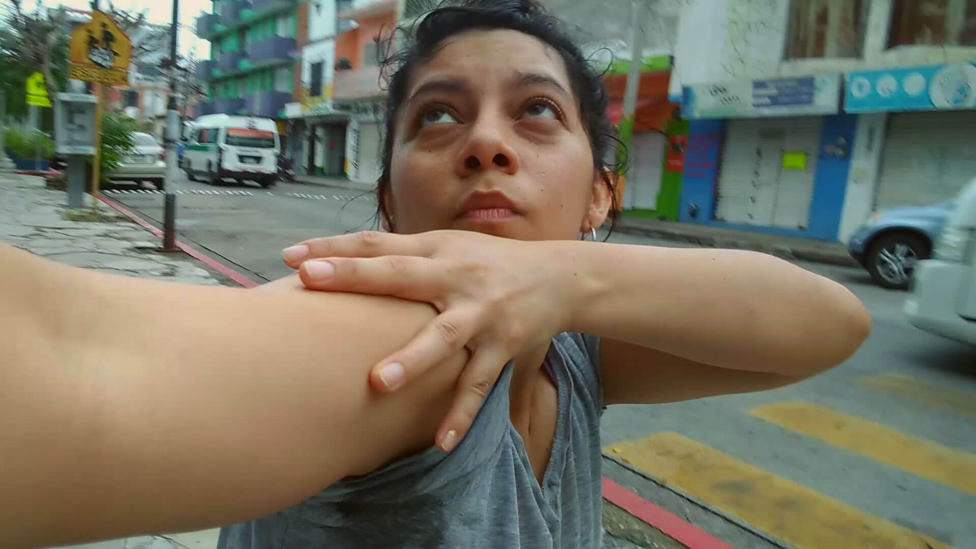 A woman with dark haired pulled away from her face, in short sleeves looks up and traces her extended right arm with left hand on a city street.