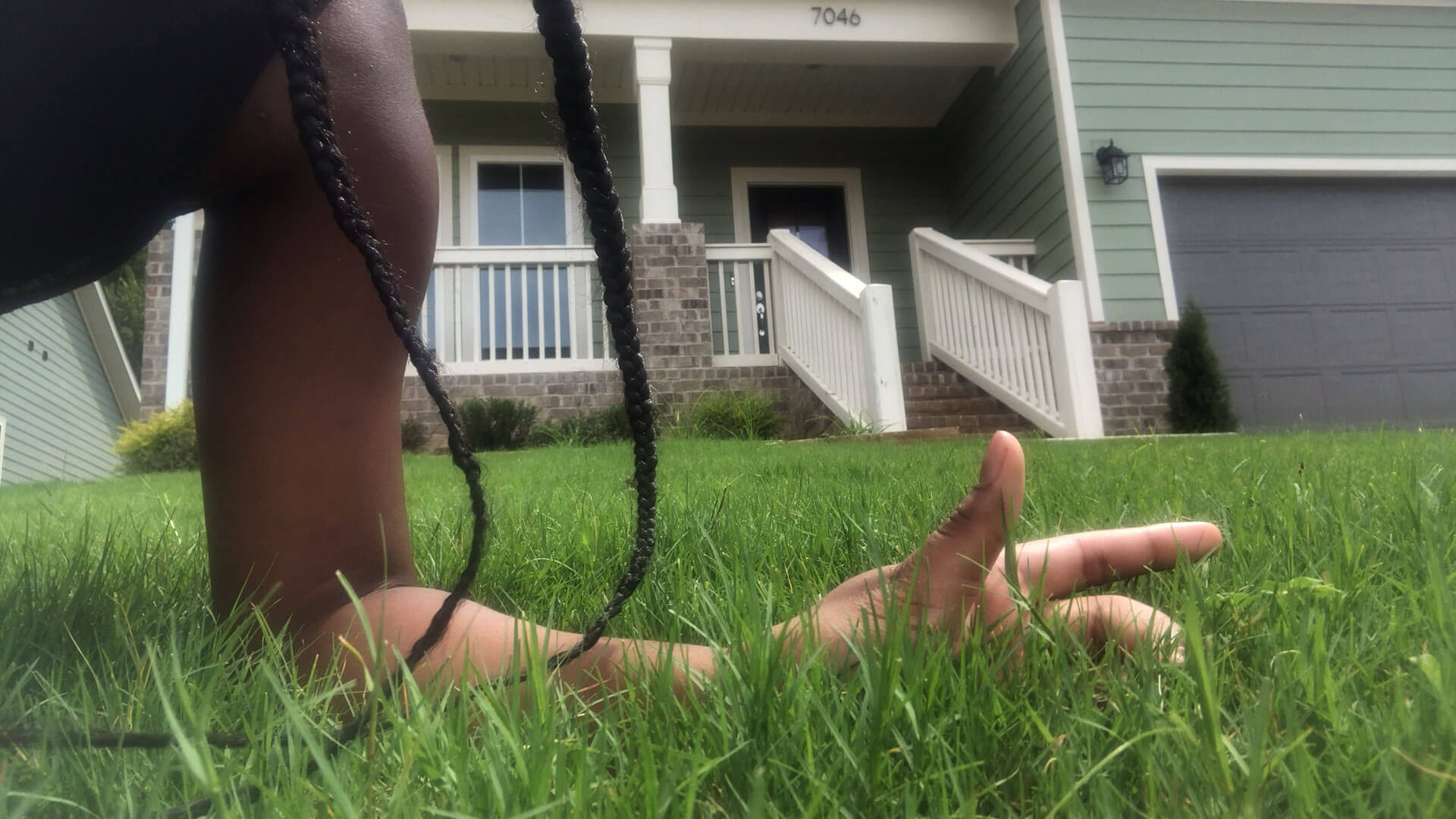 Picture of Black female of her left arm in 90 angle with forearm resting on bright green grass. Two dark braids fall to the grass, draping over her arm. In the background is a picture of a house with a white porch.