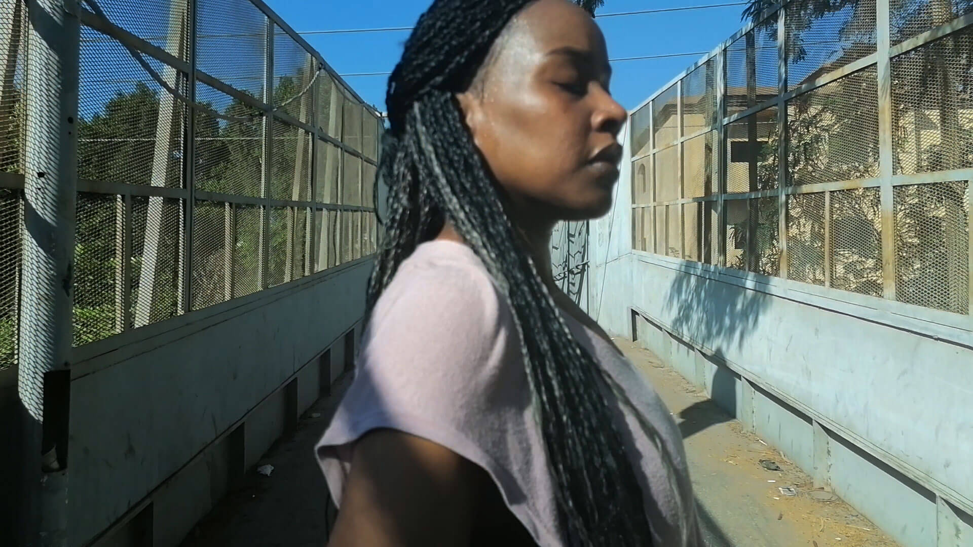 A black woman with long braids stands in profile with her eyes closed on a bridge, wearing a pink t-shirt against a blue sky.