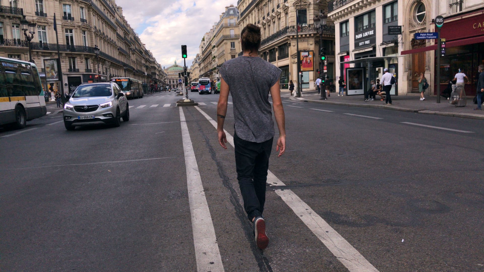 A man walks between two narrow white lines in the middle of a city street. He is wearing a grey shirt and black pants with his hair pulled in a high bun. His back is towards the camera, and to his left is a car and bus passing by.