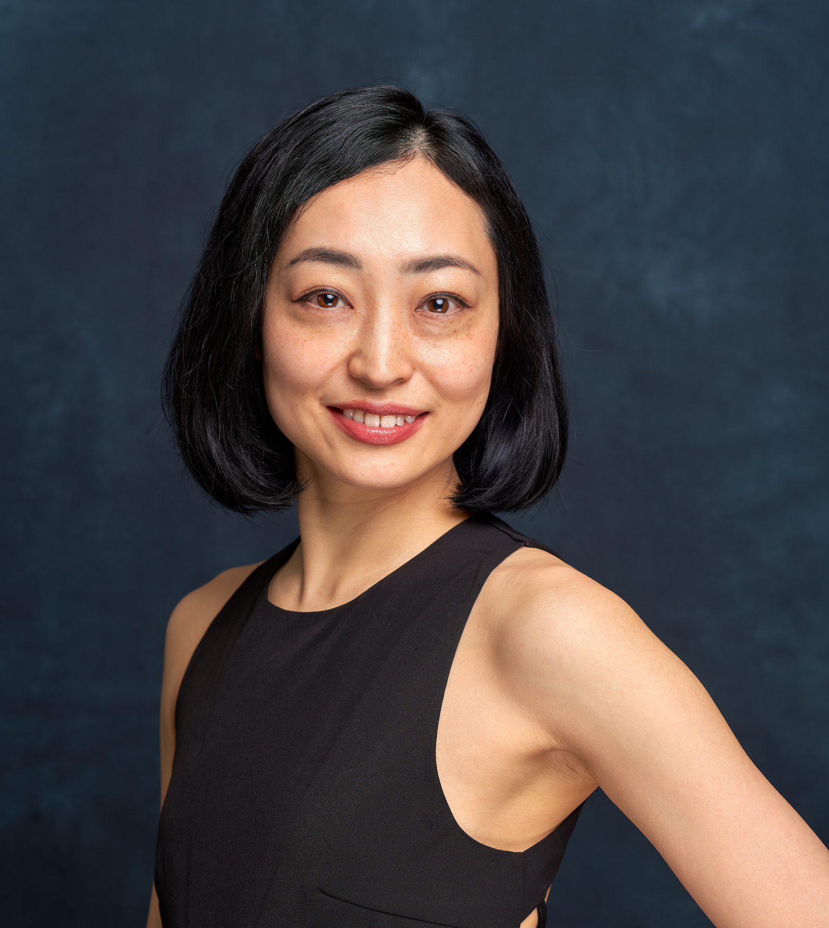 Headshot of Japanese woman smiling, wearing a black tank top and looking at camera. She has short black hair and is smiling while looking directly at camera.